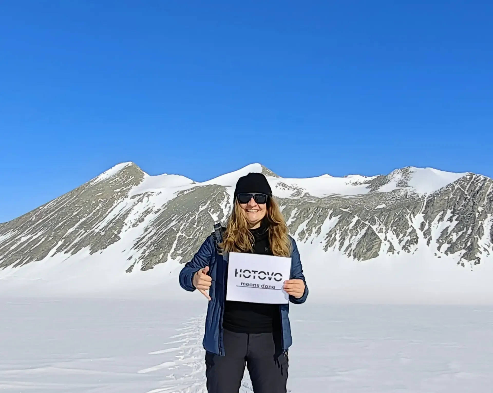 Dr. Michaela Musilova on her Astro Seven Summits mission. Holding Hotovo means done sign in her hands. Standing on thw snow, in front of the mountain
